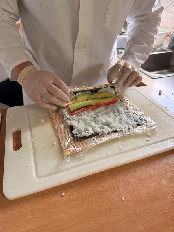 Eine Person mit weißem Kittel und Einweghandschuhen bereitet auf einem Schneidebrett Sushi zu. Auf einer Bambusmatte liegt eine Schicht Reis, darauf sind längliche Stücke von Avocado und rotem Gemüse platziert. Die Hände der Person richten die Zutaten aus.