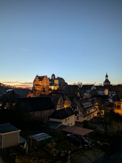 Blick über eine beleuchtete Altstadt mit Fachwerkhäusern und Burganlage auf einem Hügel bei Sonnenuntergang.