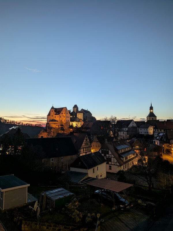 Blick über eine beleuchtete Altstadt mit Fachwerkhäusern und Burganlage auf einem Hügel bei Sonnenuntergang.