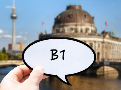Eine Hand hält eine Sprechblase mit der Aufschrift 'B1' in den Vordergrund. Im Hintergrund sind der Berliner Fernsehturm und das Bode-Museum auf der Museumsinsel in Berlin zu sehen. Der Himmel ist klar und blau, und die Architektur der Gebäude ist deutlich erkennbar.