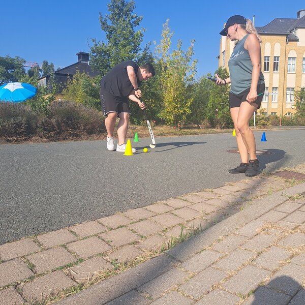 Ein Schüler führt eine kleinen gelben Ball mit einem Hockeyschläger um kleine, bunte Pylonen herum. Eine Frau mit einem Handy und einem Stift in der Hand schaut ihm dabei zu.