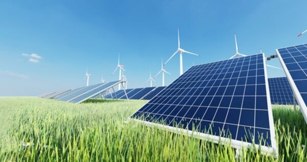 Solarzellen stehen in einem Feld mit grünen Gräsern, dahinter erkennt man mehrere moderne Windräder. Der blaue Himmel ist fast wolkenlos.