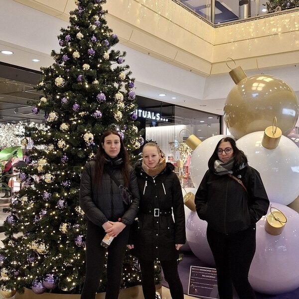 Drei Teilnehmende stehen vor großem, festlich geschmückten Weihnachtsbaum in einem Enikaufscenter.