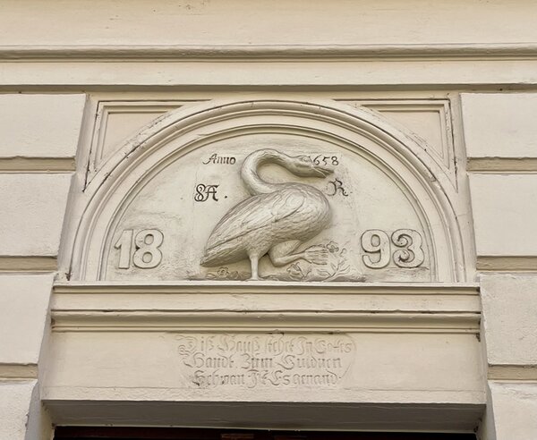 Relief eines stehenden Vogels mit langem Hals und Schnabel, vermutlich ein Schwan, in einem halbkreisförmigen Feld über einer Tür. Links und rechts sind die Zahlen '18' und '93' zu sehen, darüber steht 'Anno 1893'. Das Relief ist von dekorativen Linien und Schriftzügen umgeben, die teilweise schwer lesbar sind. Die Fassade ist hell und schlicht gehalten.