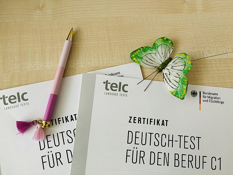 80% - Top Ergebnisse! Auf einem hellen Holztisch lieben zwei Mappen mit Deutsch-Test für den Beruf C1 in schwarz geschrieben. Links liegt ein rosa Kugelschreiber.