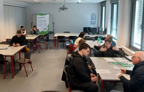 Blick in einen hellen Seminarraum, in dem mehrere Gruppen an Tischen sitzen und sich austauschen; im Hintergrund steht ein Roll-up der DIAS GmbH.