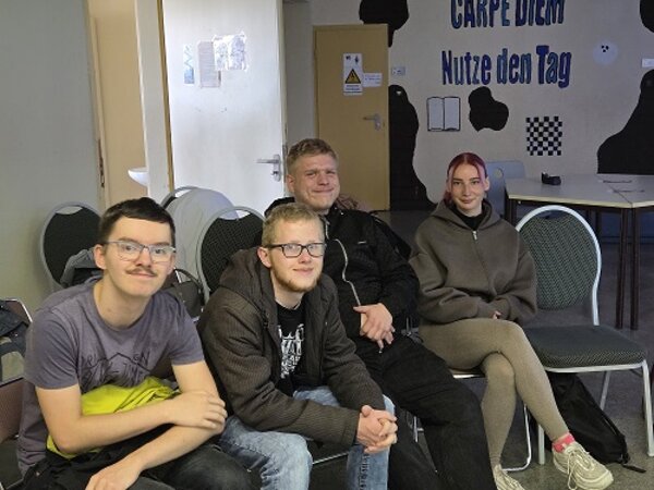 Vier junge Teilnehmende sitzen in einem Raum zusammen und blicken in die Kamera; an der Wand steht der Schriftzug „Carpe Diem – Nutze den Tag“.