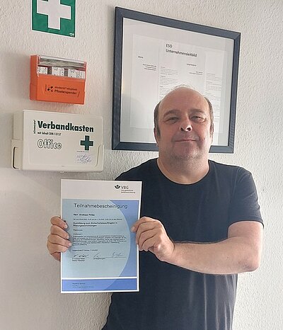 Herr Andreas Pötke zum Sicherheitsbeauftragten für Bildungseinrichtungen (SIB) weitergebildet Ein Mann in schwarzem T-Shirt steht vor einer weißen Wand mit Erste-Hilfe Kasten und hält ein hellblaues Zertifikat in die Kamera.