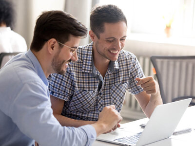 Zwei lächelnde Männer sitzen nebeneinander an einem Tisch und arbeiten gemeinsam an einem Laptop. Der Mann links trägt ein hellblaues Hemd und eine Brille, während der Mann rechts ein kariertes Hemd trägt. Beide wirken begeistert und konzentriert. Der Mann rechts zeigt mit dem Finger auf den Bildschirm, während der Mann links ihm über die Schulter schaut. Der Hintergrund zeigt einen hellen Raum, möglicherweise ein Büro oder ein Besprechungszimmer.