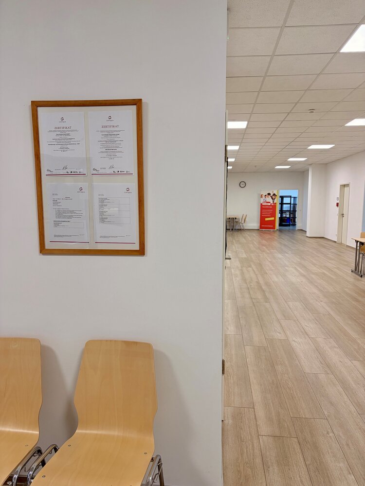 Flur- und Wartebereich der Schule mit Sitzgelegenheiten, Zertifikaten an der Wand und Blick in helle Unterrichtsräume.