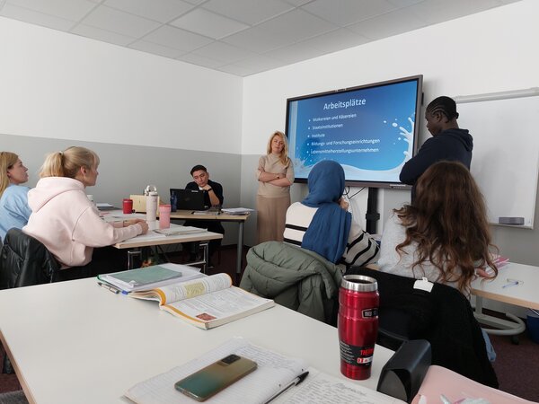 Eine Frau und ein Mann stehen vor einem Bildschirm mit Informationen zu verschiedenen Arbeitsplätzen. Mehrere Teilnehmer, darunter Frauen mit Kopftüchern, sitzen an den Tischen und hören zu.