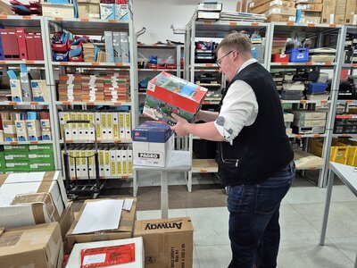 Ein Auszubildender beim Stapeln und Prüfen von Kartons im Lagerregal, umgeben von Aktenordnern und Büroartikeln.