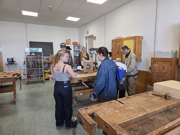 In einem Raum mit Tischlerbänken und Werkzeug stehen mehrere Erwachsene und Jugendliche.