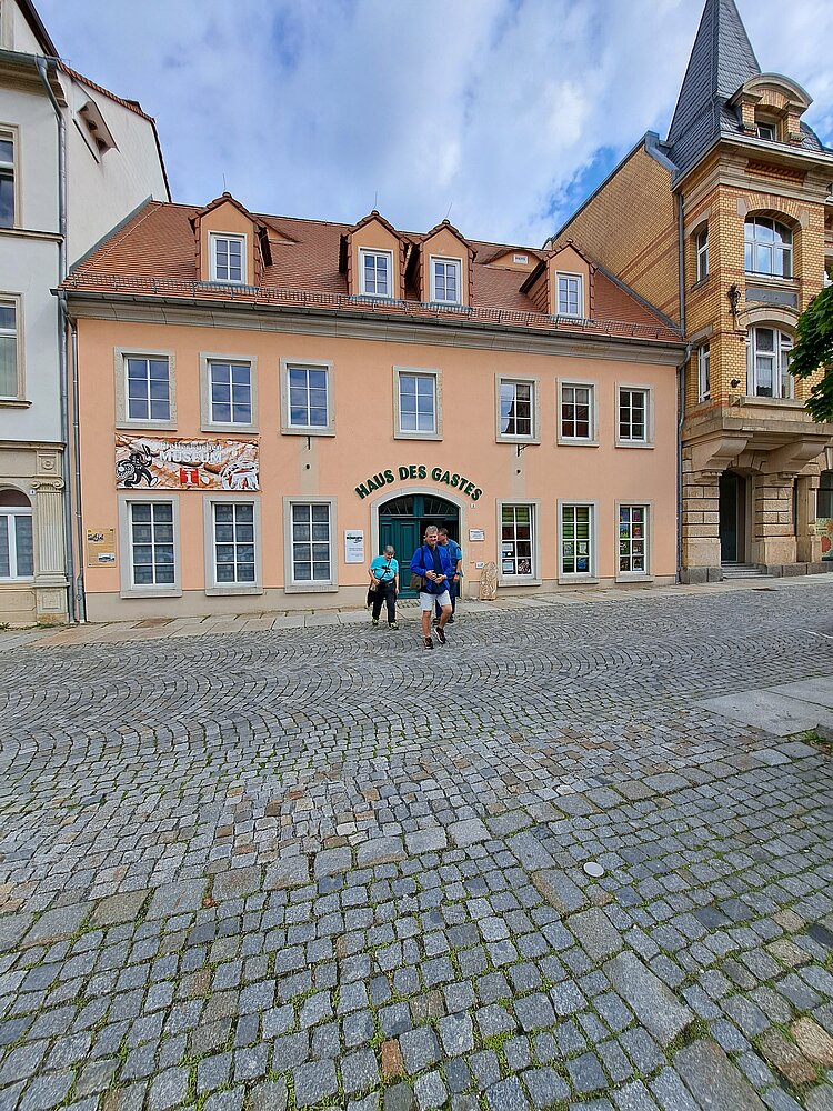 Außenansicht des Gebäudes „Haus des Gastes“ mit Kopfsteinpflaster im Vordergrund. Drei Personen stehen vor dem Eingang, das Gebäude hat eine rosa Fassade mit mehreren Fenstern und einem roten Dach.