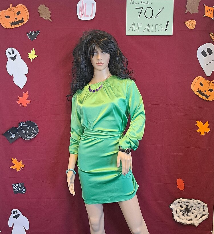 BRAFO-Projekt im Oktober – Herbst trifft Halloween! Eine große weibliche Schaufensterpuppe mit dunklen, langen Haaren trägt ein hellgrünes Kleid. Im Hintergrund kleben auf einem dunkelroten Laken selbst gebastelte Halloween-Dekorationen wie Kürbisse, Geister, Fledermäuse und Spinnennetze.