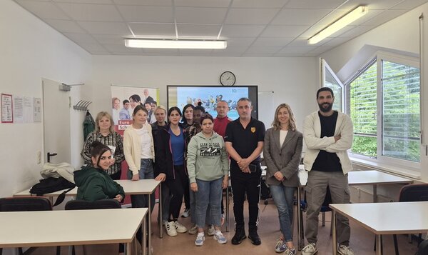 In einem hellen Klassenraum stehen mehrere Teilnehmende in lockerer Formation zwischen Tischen und posieren für ein Foto. Im Hintergrund ist ein Bildschirm mit einer Uhrzeit zu sehen, daneben ein aufgestelltes Plakat mit mehreren Porträts. Die Fenster auf der rechten Seite sind weit geöffnet, draußen ist grüne Vegetation sichtbar. Die Personen blicken in Richtung Kamera, einige lächeln, andere haben einen neutralen Gesichtsausdruck. Die Kleidung ist überwiegend leger, eine Person trägt einen dunklen Pullover mit einem auffälligen Emblem auf der Brust.