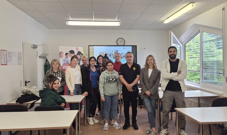In einem hellen Klassenraum stehen mehrere Teilnehmende in lockerer Formation zwischen Tischen und posieren für ein Foto. Im Hintergrund ist ein Bildschirm mit einer Uhrzeit zu sehen, daneben ein aufgestelltes Plakat mit mehreren Porträts. Die Fenster auf der rechten Seite sind weit geöffnet, draußen ist grüne Vegetation sichtbar. Die Personen blicken in Richtung Kamera, einige lächeln, andere haben einen neutralen Gesichtsausdruck. Die Kleidung ist überwiegend leger, eine Person trägt einen dunklen Pullover mit einem auffälligen Emblem auf der Brust.