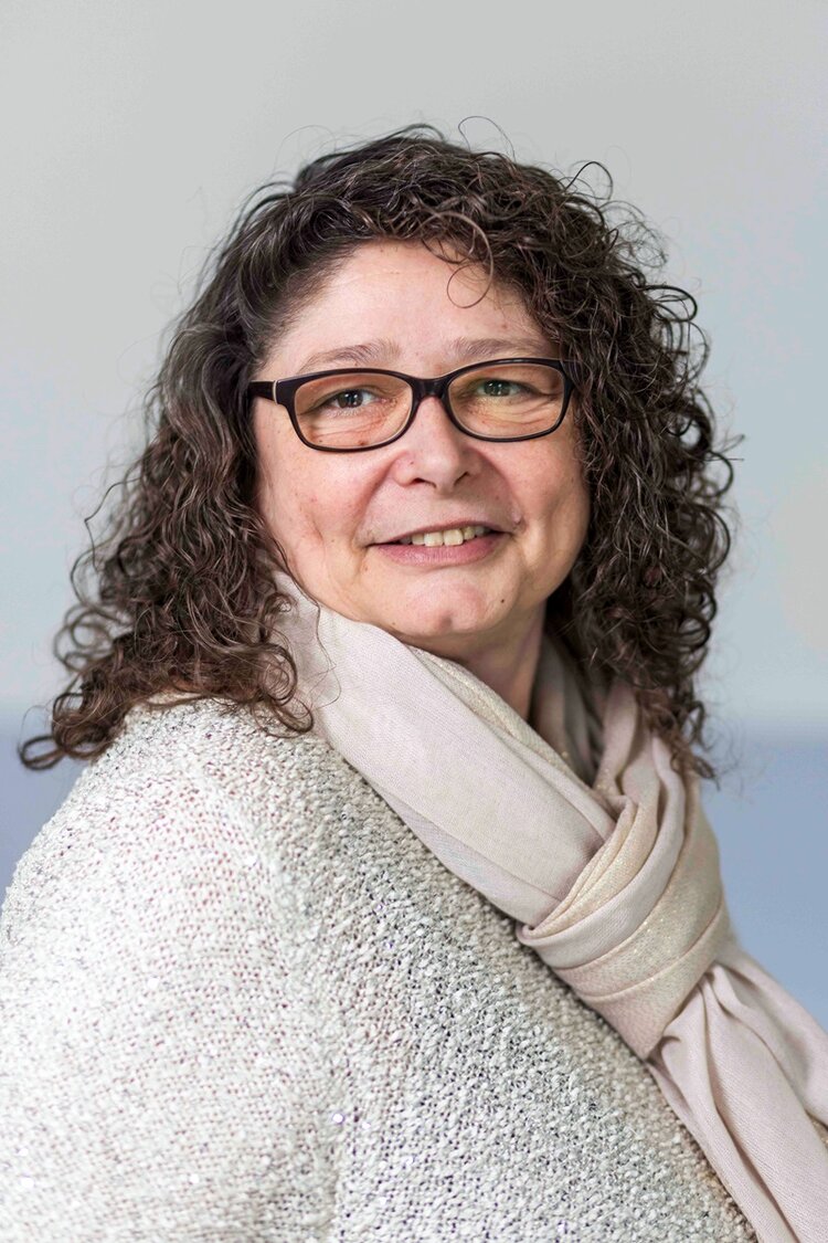 Eine weibliche Person mit lockigem, braunem Haar trägt einen hellen Schal und einen cremefarbenen, strukturierten Pullover und lächelt für ein Porträtfoto in die Kamera. Der Hintergrund ist einfarbig und hell.