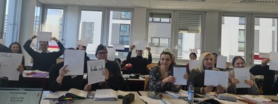 Eine Gruppe von jungen Frauen und Männern sitzen in zwei Tischreihen zur Kamera gewendet und halten ausgedruckte Texte und handschriftlich angefertigte Notizen hoch.