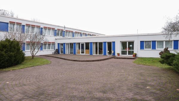 Innenhof eines zweigeschossigen, modernen Gebäudekomplexes mit weißer Fassade und blauen Fensterrahmen; im Vordergrund ein kreisförmig gepflasterter Platz, umgeben von Rasenflächen, Sträuchern und kahlen Bäumen, im Hintergrund große Fensterfronten und Eingänge.