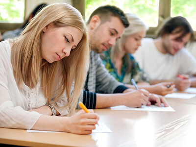 Eine Gruppe von jungen Erwachsenen sitzt konzentriert in einem hellen Seminarraum und schreibt Notizen auf Blätter Papier. Im Vordergrund ist eine junge Frau mit langen blonden Haaren zu sehen, die nachdenklich auf ihr Blatt schaut, während sie mit einem gelben Stift schreibt. Im Hintergrund sind weitere Teilnehmer:innen zu erkennen, die ebenfalls in ihre Aufgaben vertieft sind. Der Raum wirkt freundlich und einladend durch das natürliche Licht, das durch die großen Fenster fällt.
