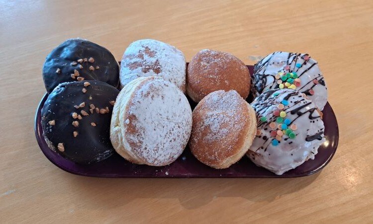 Sechs verschiedene Berliner (Pfannkuchen, Kreppel, Krapfen) auf einem länglichen, dunklen Teller: zwei mit Schokoladenglasur und Nüssen, zwei mit Puderzucker bestäubt, einer mit einfacher Zuckerglasur und einer mit bunter Streuselglasur.