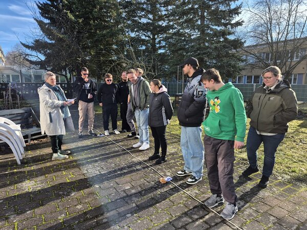 Draußen steht eine Gruppe Teilnehmender im Halbkreis auf einem gepflasterten Hof; eine Person liest von einem Blatt vor, auf dem Boden liegt eine gespannte Schnur als Teil einer Übung.