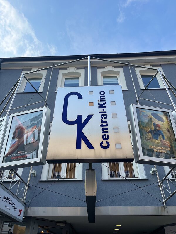Eine blaue Hauswand mit einer vorgebauten Metallkonstruktion. Daran hängt der Schriftzug des Kinos "CK Central Kino" und links und rechts davon jeweils ein Plakat von aktuellen Filmen.