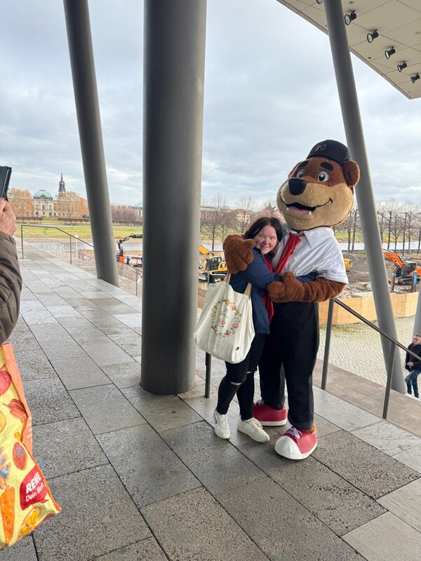 Junge Frau umarmt ein Bären-Maskottchen auf dem Vorplatz; Säulen und Stadtlandschaft im Hintergrund, freundliche Szene.