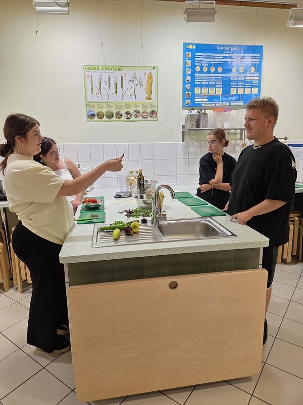 Eine Gruppe von Personen steht in einer Gemeinschaftsküche und unterhält sich. Auf der Spüle zwischen ihnen liegt Gemüse. Im Hintergrund sind Poster an der Wand zu sehen, darunter eines mit dem Titel 'Käseherstellung'.