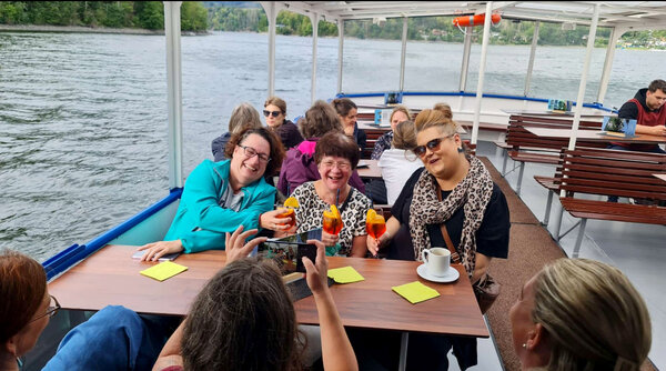 Drei Frauen sitzen während einer Schifffahrt an einem Tisch auf dem Außendeck, lachen und stoßen mit orangefarbenen Getränken an.