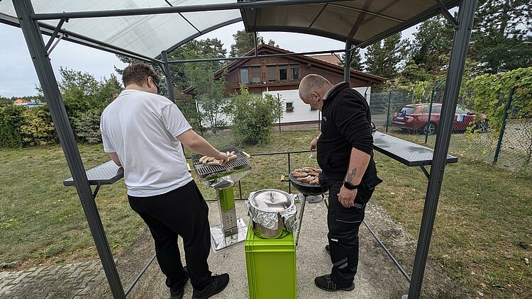 Rückblick Sportfest 2024 Leibliches Wohl. Zwei Männer grillen im Garten der Euro-Schulen Hoyerswerda mit zwei Kugelgrills Würstchen.