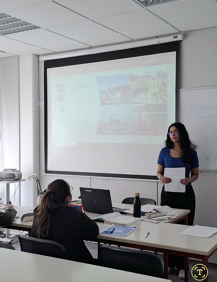 Eine weibliche Teilnehmerin mit langen dunklen Haaren und einer Brille im blauen Oberteil steht vorn am Whiteboard im Unterrichtsraum und hält ihre Präsentation vor ihren Mitteilnehmenden des B2-Sprachkurses an den Euro-Schulen Magdeburg.
