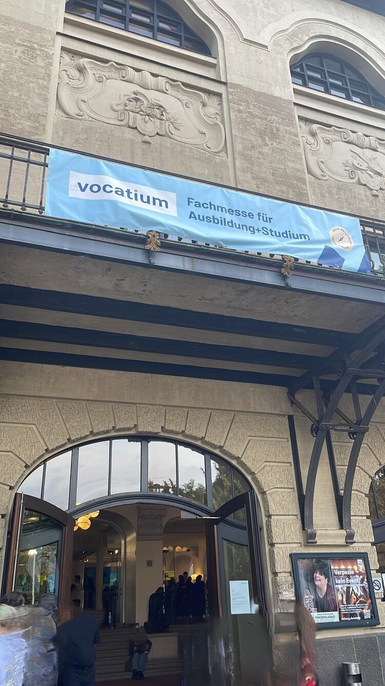 Der Eingang eines historischen Gebäudes mit der Aufschrift „vocatium Fachmesse für Ausbildung+Studium“ auf einem blauen Banner. Über dem Eingang sind verzierte Steinmetzarbeiten zu sehen. Menschen betreten das Gebäude durch eine große, offene Glastür. Das Gebäude wirkt einladend und ist vermutlich ein Veranstaltungsort für Bildungsmessen.