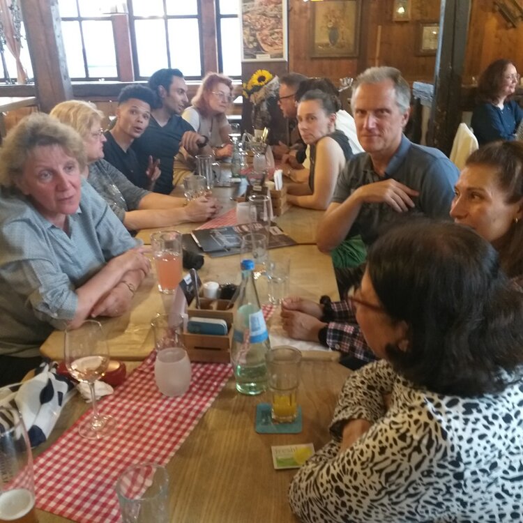 Die Gruppe der Mitarbeitenden sitzt an einem langen Holztisch in einem rustikalen Restaurant. Auf dem Tisch stehen verschiedene Gläser, Flaschen und Teller, darunter eine Flasche Mineralwasser, Weingläser und ein Glas mit rosa Getränk. In der Mitte des Tisches liegt eine rot-weiß karierte Stoffserviette. Die Personen sind in angeregter Unterhaltung, einige schauen in Richtung Kamera. Im Hintergrund sind Holzvertäfelungen, Fenster und Wandbilder zu sehen. Die Atmosphäre wirkt gesellig und entspannt.