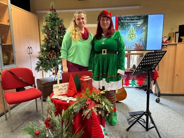 Zwei Frauen vor weihnachtlich dekoriertem Lesepult, links ein Weihnachtsbaum, rechts eine Leinwand mit dem Buchcover „Wichtelweihnacht“.