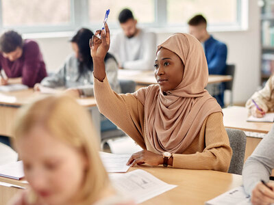 Eine Frau mit einem beigen Hijab und einem gestreiften Pullover sitzt in einem Klassenzimmer und hebt ihre Hand, um eine Frage zu stellen oder eine Antwort zu geben. Sie trägt eine Armbanduhr und hält einen Stift in der Hand. Im Hintergrund sind andere Schüler zu sehen, die an ihren Tischen sitzen und arbeiten. Der Raum ist hell und hat große Fenster, die Tageslicht hereinlassen.