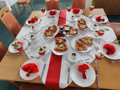 Eine Nahaufnahme des gedeckten Tisches mit herzförmigen Pizzen, Gläsern mit roten Getränken und kleinen Desserts in Gläsern. Der Tisch ist mit roten Servietten und einer roten Tischdecke dekoriert.