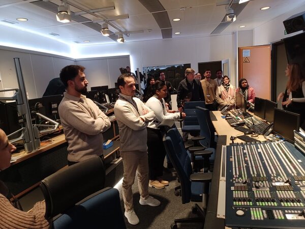 Eine Gruppe von Personen steht in einem Technikraum vor Bürostühlen und Tischen, auf denen verschiedene Steuerungspulte und Monitore stehen. Es handelt sich um den Regieraum eines Fernsehstudios. Die Personen hören einer Frau zu, die vor ihnen steht und spricht.