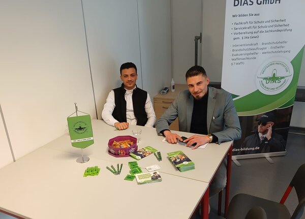 Zwei Vertreter der DIAS GmbH sitzen an einem Infotisch mit Flyern, Kugelschreibern und Werbematerialien vor einem Roll-up-Banner.