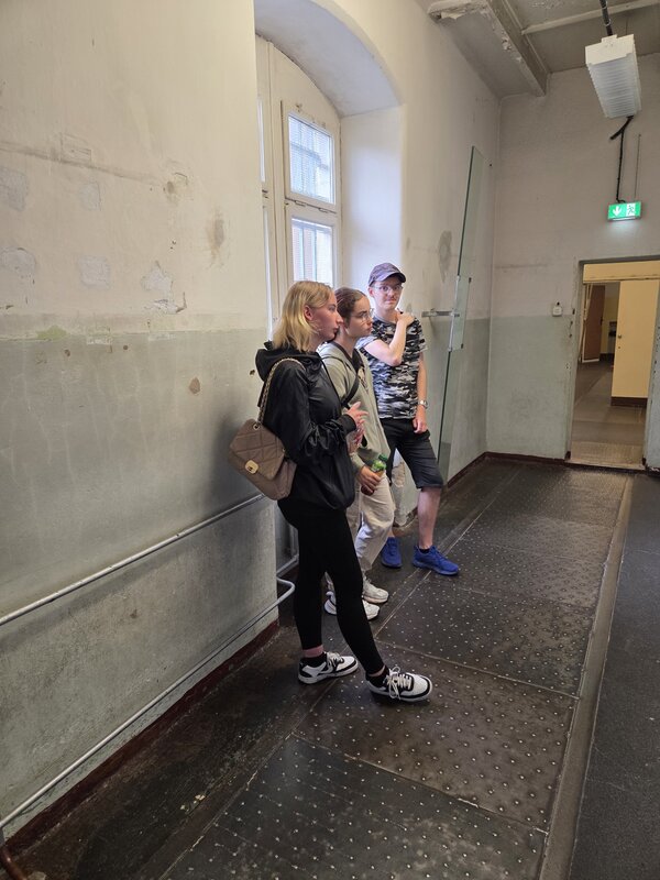Drei junge Menschen stehen in einem alten, kahlen Flur mit hohen Fenstern. Die Wände sind teilweise beschädigt, die Atmosphäre wirkt kühl und historisch.