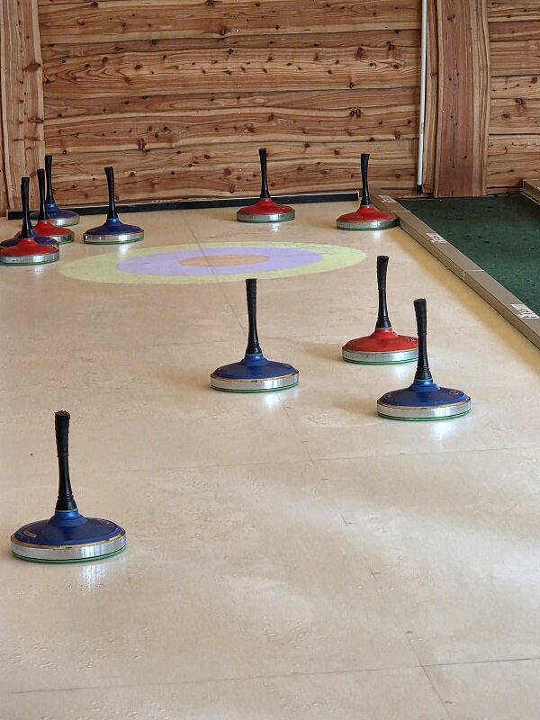 Mehrere Eisstock-Spielsteine liegen auf einer glatten Bahn, im Hintergrund eine hölzerne Wand und eine Zielmarkierung auf dem Boden