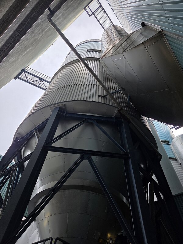 Blick von unten auf mehrere hohe, zylindrische Industrieanlagen aus Metall. Die Behälter und Rohrleitungen sind von Stahlkonstruktionen, Laufstegen und großen Verkleidungen umgeben. Im Hintergrund ist der bewölkte Himmel zwischen den Gebäudeteilen sichtbar.