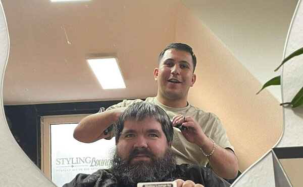Blick in einen Spiegel: Ein Friseur schneidet einem Kunden mit langem, graumeliertem Bart die Haare. Im Hintergrund ist ein Spiegel zu sehen, in dem ein Teil des Friseursalons mit der Aufschrift "STYLING Lounge".