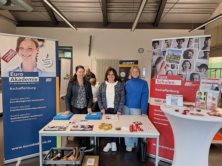 Auf der Weiterbildungsmesse Main.Fortschritt Frau Beller, Frau Crétien und Frau May strahlen am Messestand neben dem Roll-up der Euro-Schulen um die Wette in die Kamera. Auch einige Messegeschenke sind auf den Tischen zu sehen.