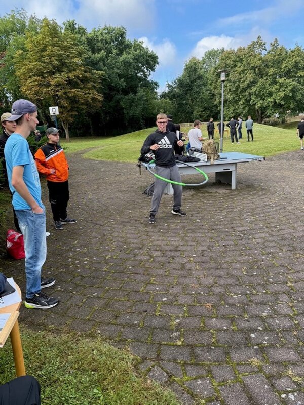 Im Mittelpunkt steht eine Person mit Brille und dunklem Trainingsanzug, die einen Hula-Hoop-Reifen um die Hüfte kreisen lässt. Die Szene spielt sich im Freien auf einer gepflasterten Fläche ab. Im Hintergrund stehen weitere Personen, einige an einem Tischtennistisch, andere auf einer Wiese. Links im Bild beobachten mehrere Menschen das Geschehen, einer trägt ein hellblaues T-Shirt und eine Kappe. Im Hintergrund sind Bäume und ein Basketballkorb zu sehen. Der Himmel ist teilweise bewölkt.