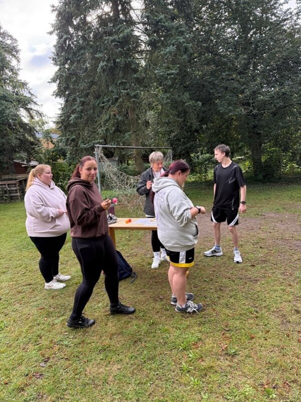 Fünf Personen stehen auf einer Wiese vor einem Holztisch. Sie tragen sportliche Freizeitkleidung wie Shorts, Trainingshosen und Kapuzenpullover. Im Hintergrund sind ein Fußballtor, Bäume und ein Stapel Holz zu sehen. Die Personen unterhalten sich oder beschäftigen sich mit Gegenständen in den Händen.