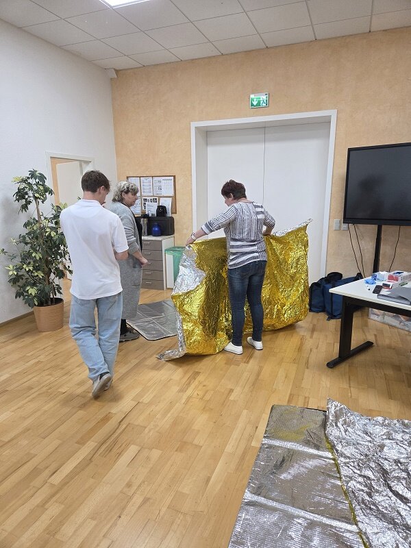 Die Mitarbeitenden der Erste-Hilfe-Fortbildung bereiten eine goldene Rettungsdecke für eine Übung vor. Im Hintergrund sind ein Bildschirm und Schulungsmaterialien, eine große Grünpflanze, ein Tisch und ein großer Bildschirm zu sehen.