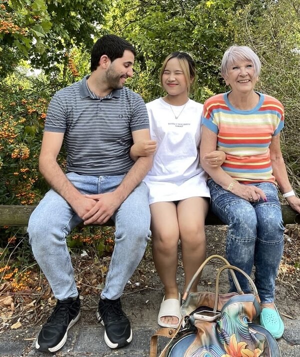 Zwei Teilnehmende sitzen mit ihrer Dozentin auf einem Holzbalken und posieren für ein Abschiedsfoto. 