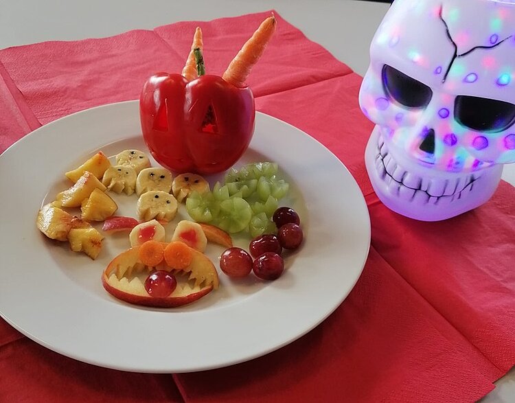 BRAFO-Projekt im Oktober – Herbst trifft Halloween! Auf einem weißen Teller liegen Obst und Gemüse, die gruselig gestaltet wurden. Darunter Bananenscheiben, die zu kleinen Geistern geschnitten wurden, eine rote Paprika, in die Augen und ein Mund geschnitz sind und in der Karotten oben drin stecken, außerdem Apfelscheiben, die wie ein zackiger Monstermund mit Karottenaugen und Traubenzunge dekoriert sind. Auf dem Tisch liegt zudem ein beleuchteter Totenkopf als Dekoration.